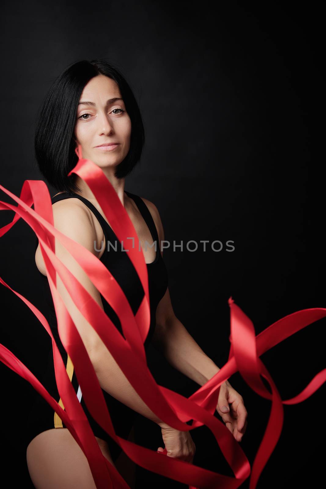 young woman gymnast of Caucasian appearance with black hair spins red satin ribbons, gymnastic exercises on black background, selective focus