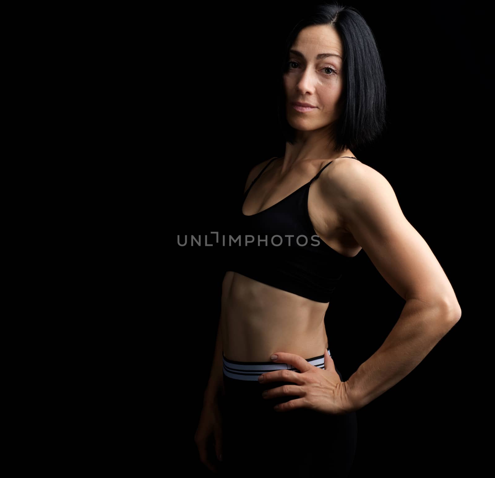 Adult girl with a sports figure in black bra and black shorts standing on a dark background, muscular body, black hair, low key