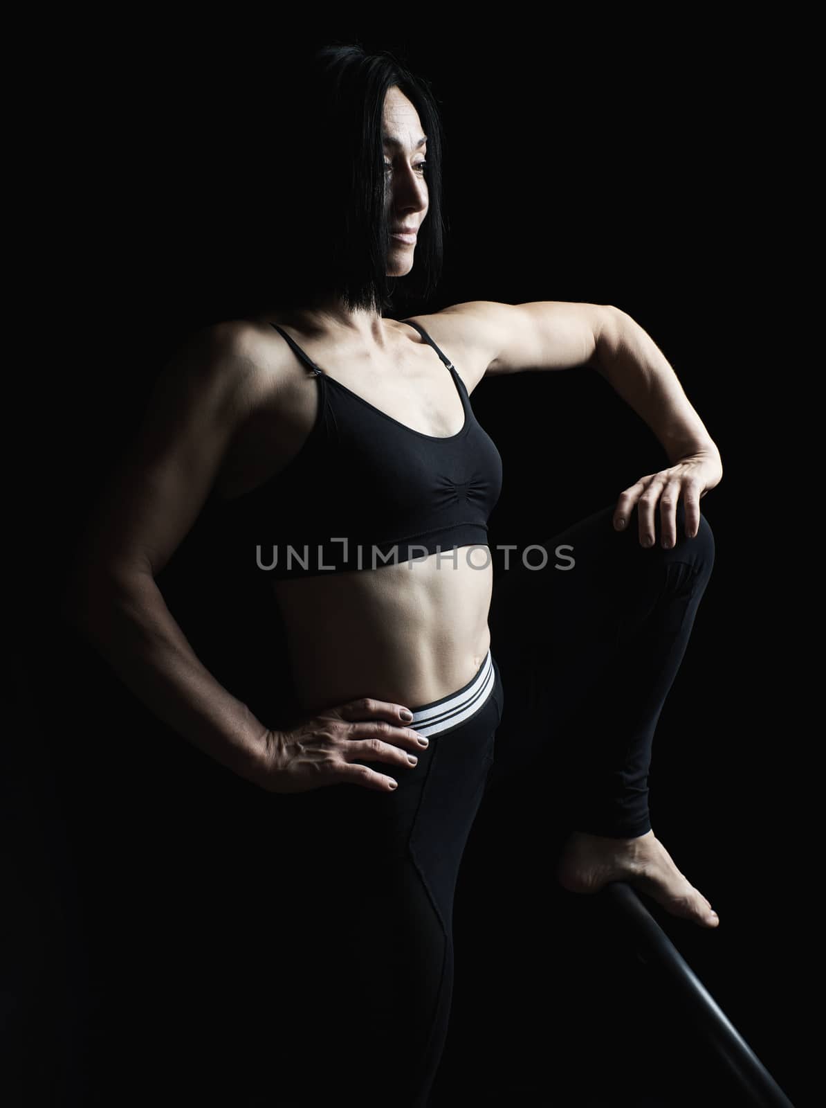 young beautiful athletic girl with muscles in black uniform posing sideways, leg raised up, studio photo in low key