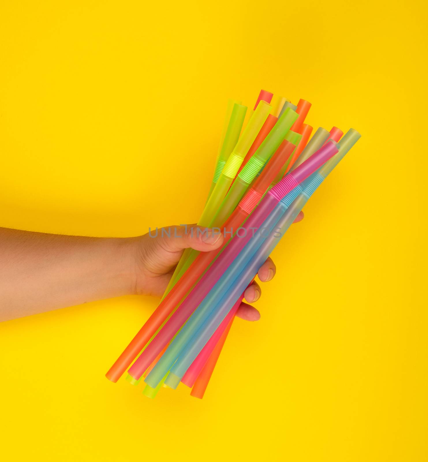 female hand holding multicolored plastic cocktail tubes, yellow background