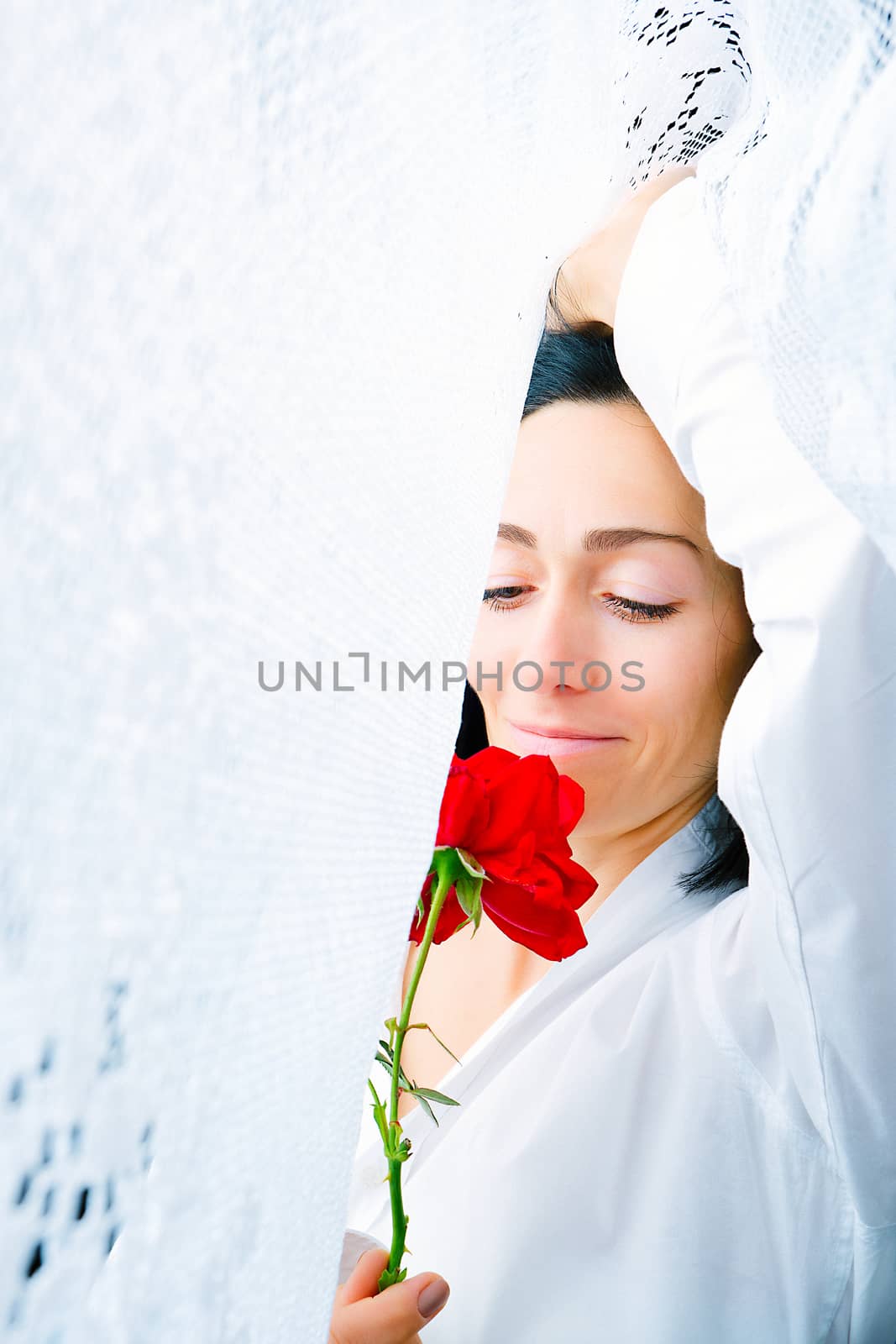 portrait of a young woman with a red rose in her hands surrounded by lace white curtains, waking up in the early morning