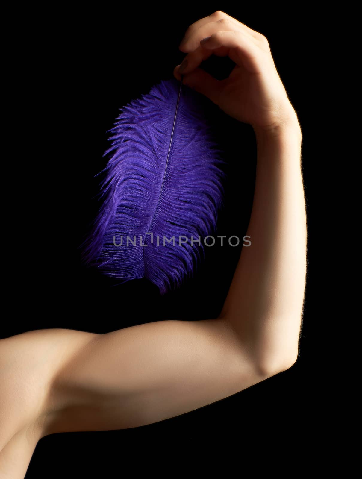 female hand is bent at the elbow, the muscles are tense and hold a blue feather on a dark background, concept of light and heavy