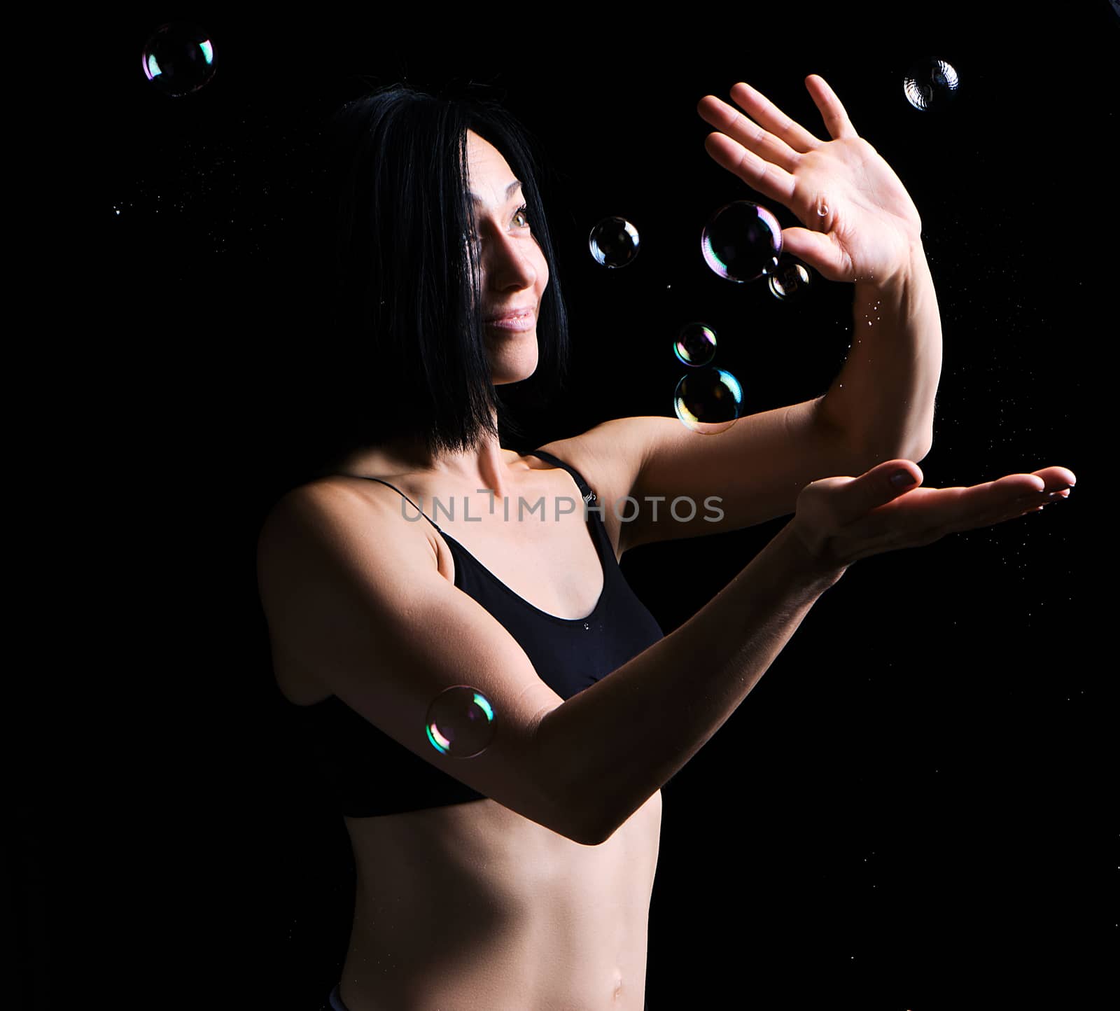 beautiful athletic woman with muscular body dressed with a black topic catches soap bubbles on a black background, studio portraits in a dark key