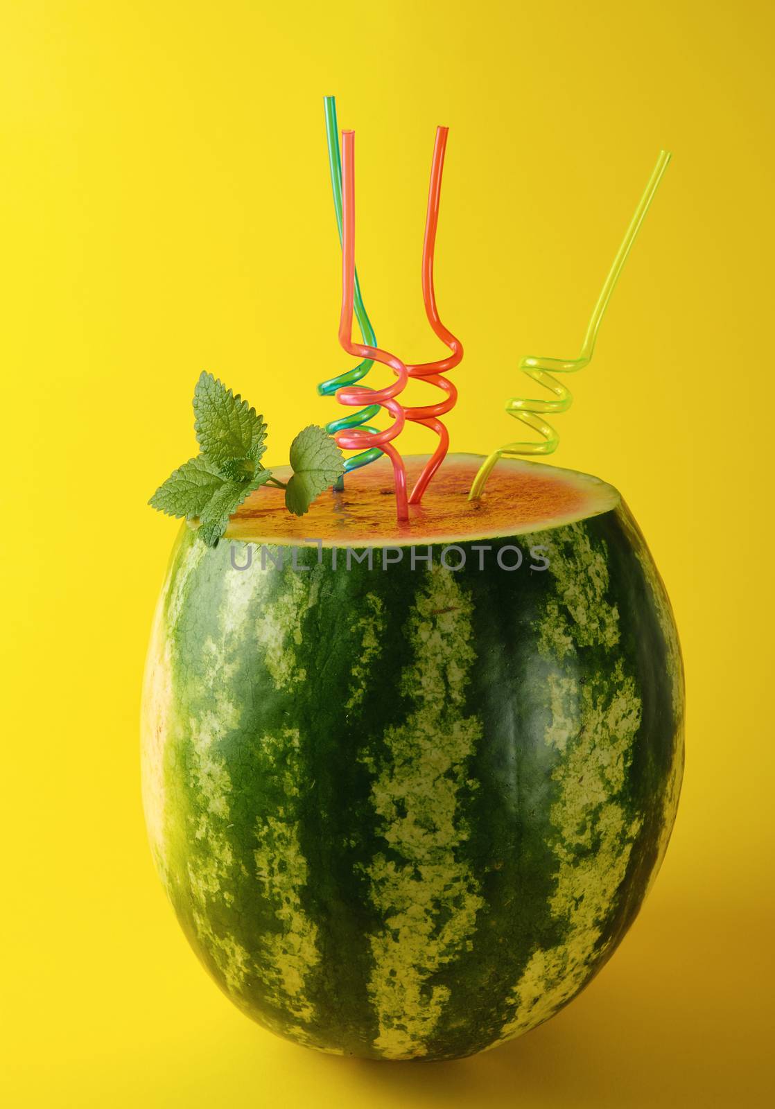 ripe striped green watermelon is cut and multicolored plastic cocktail tubes with a sprig of mint on a yellow background,  refreshing summer cocktail for a friendly company