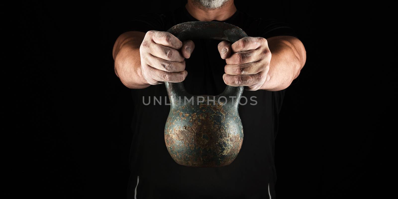 adult strong athlete in black clothes holding an iron kettlebell on his outstretched arms, black background