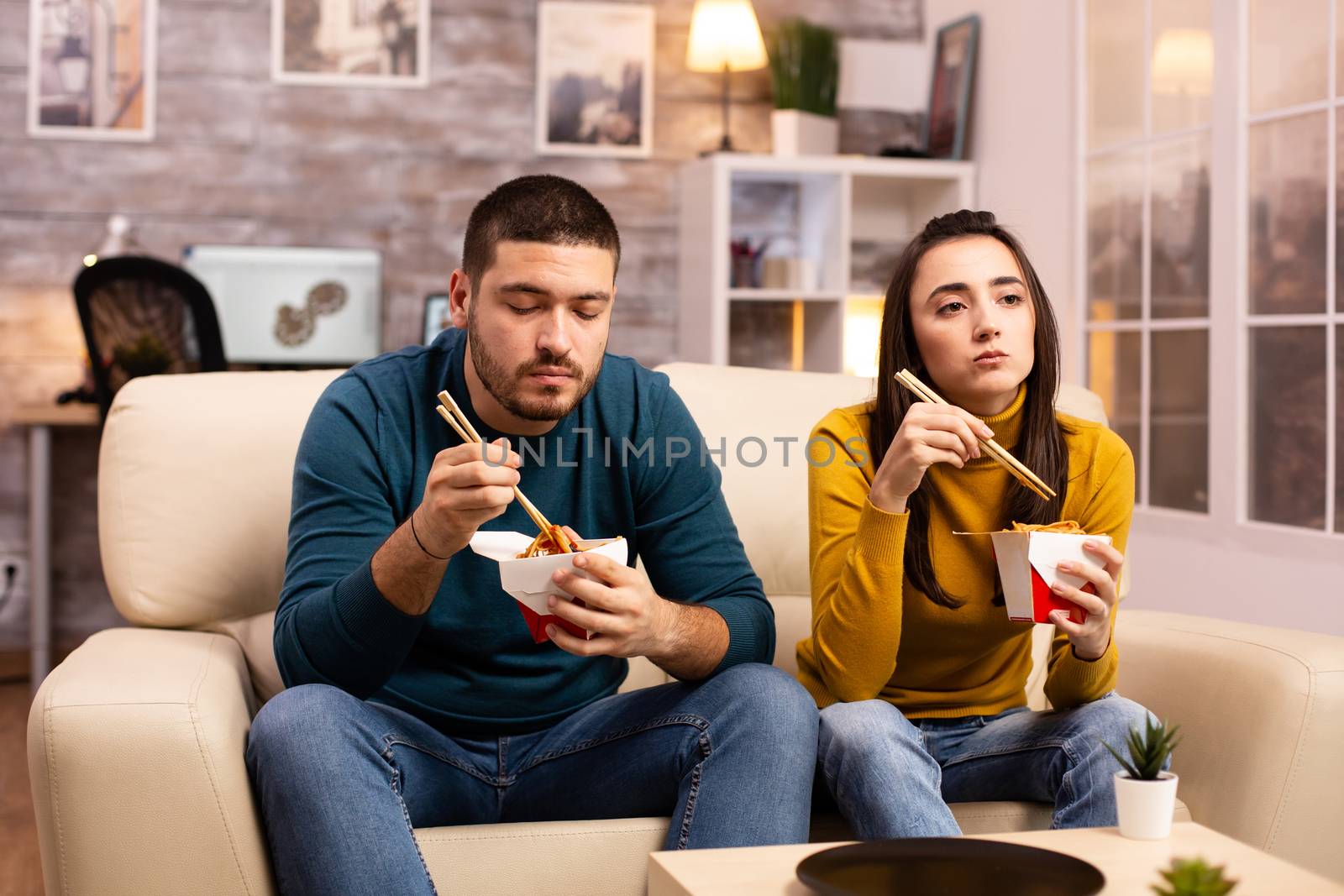 In modern cozy living room couple is enjoying takeaway noodles while watching TV by DCStudio