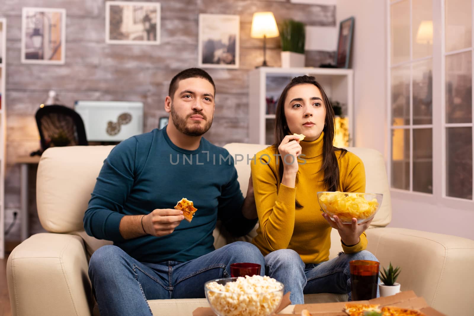 Beautiful young couple watching TV and eating fast food takeaway in the living room