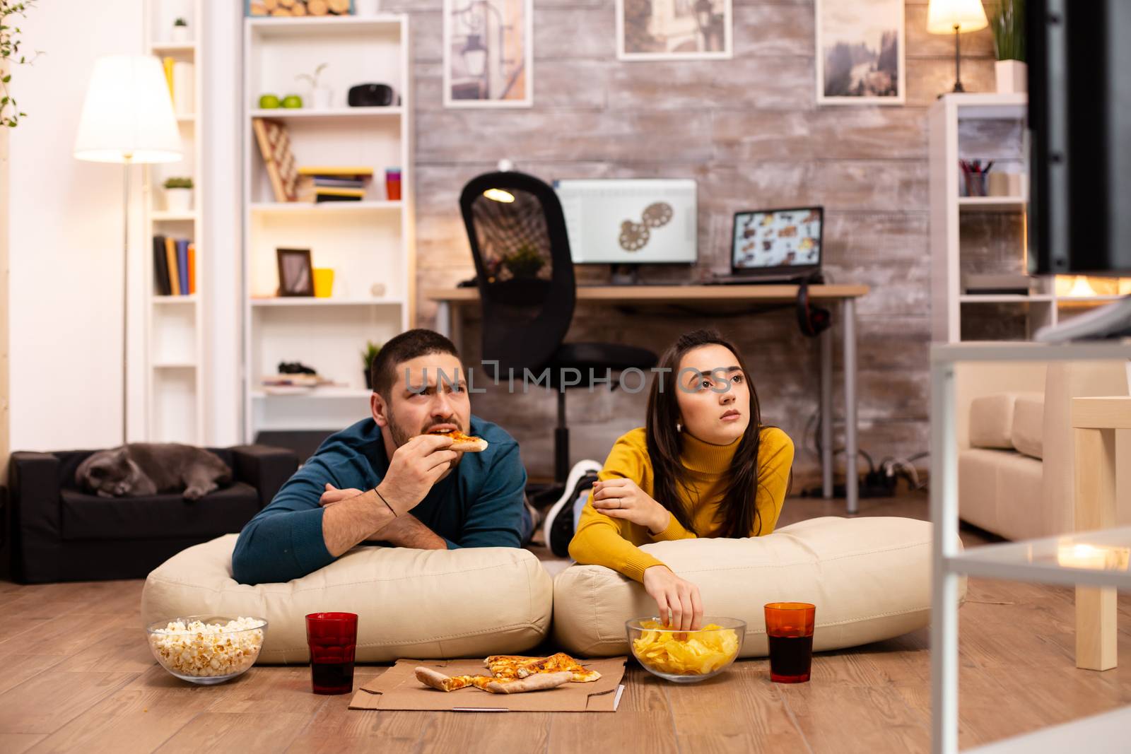 Couple sitting on the floor and watching TV in their living room by DCStudio