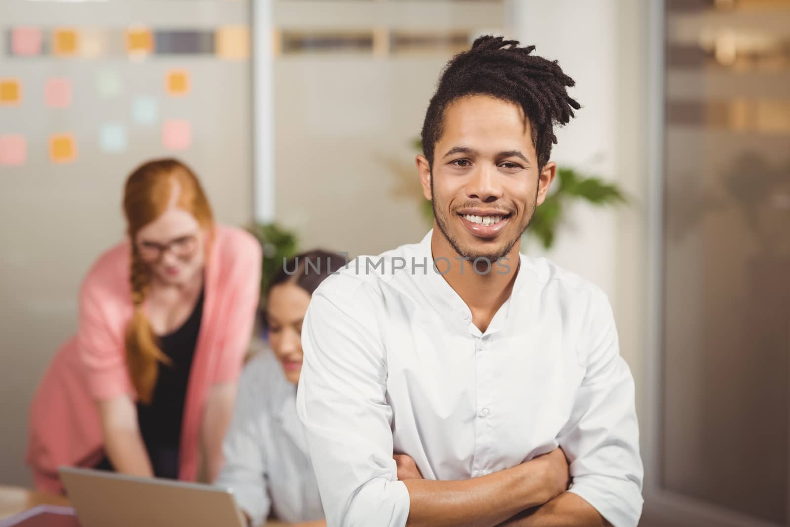 Portrait of smiling businessman with arms crossed in office with female colleagues working in background