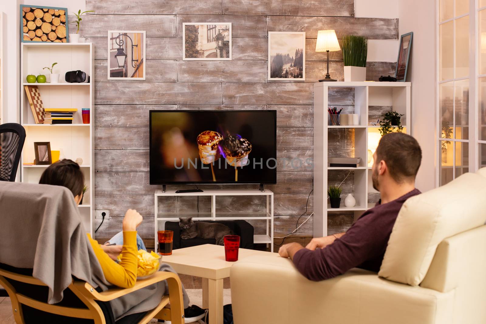 Back view of couple in living room watching a movie on the TV while eating takeaway food