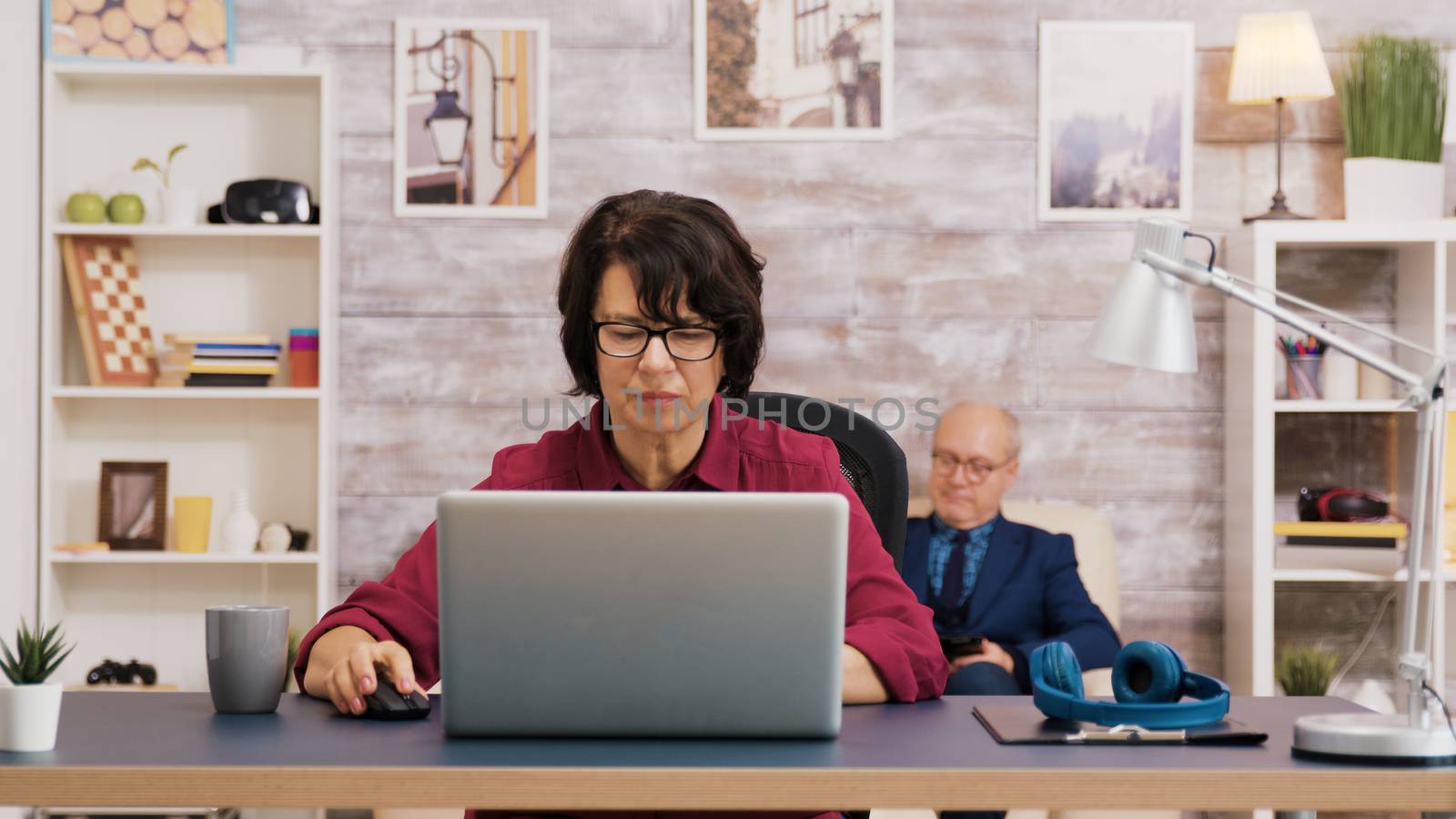 Elderly age woman taking a sip of coffee while browsing on laptop. Old man relaxing on sofa in the background.