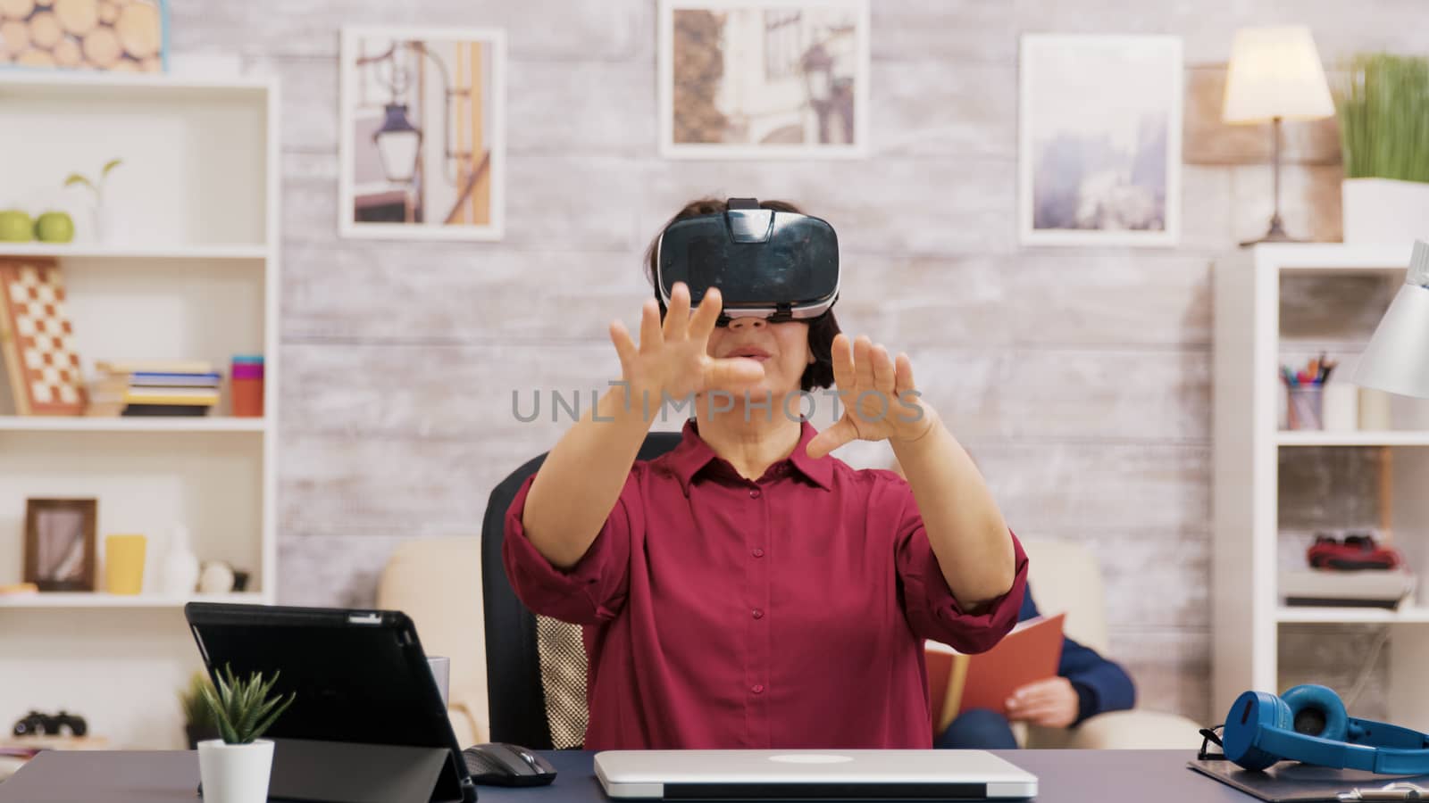 Retired senior woman amazed while using virtual reality goggles in living room. Elderly man sitting on sofa while reading a book.