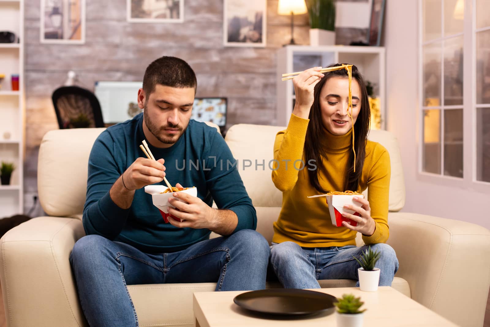 In modern cozy living room couple is enjoying takeaway noodles while watching TV comfortably on the sofa