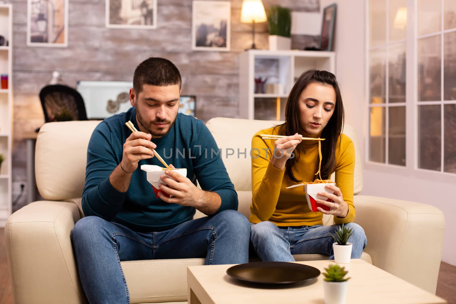 In modern cozy living room couple is enjoying takeaway noodles while watching TV comfortably on the sofa