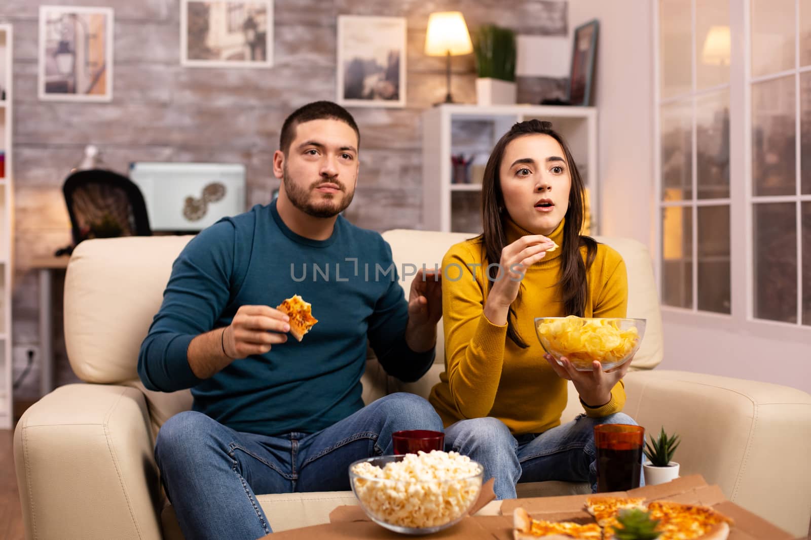 Beautiful young couple watching TV and eating fast food takeaway in the living room