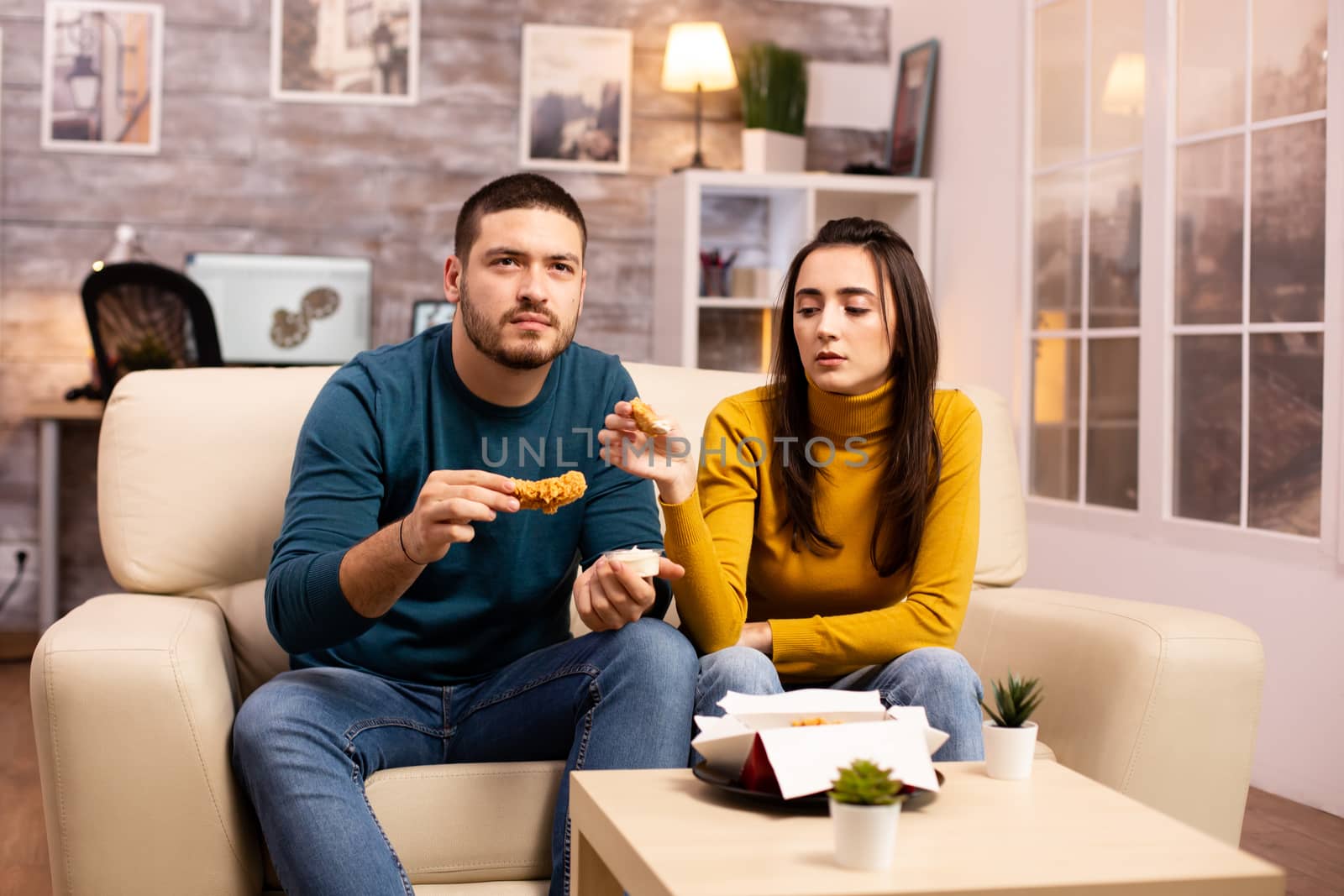 Young couple eating fried chicken in front of the TV in the living room