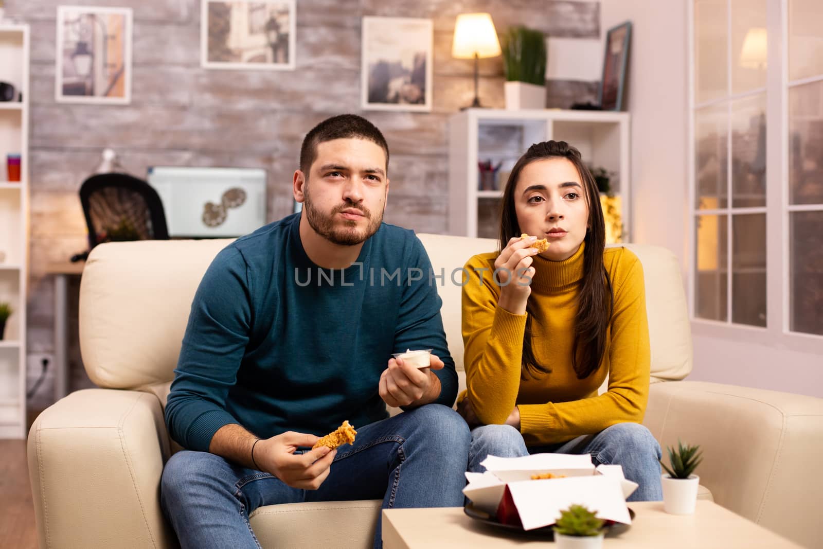 Young couple eating fried chicken in front of the TV in the living room