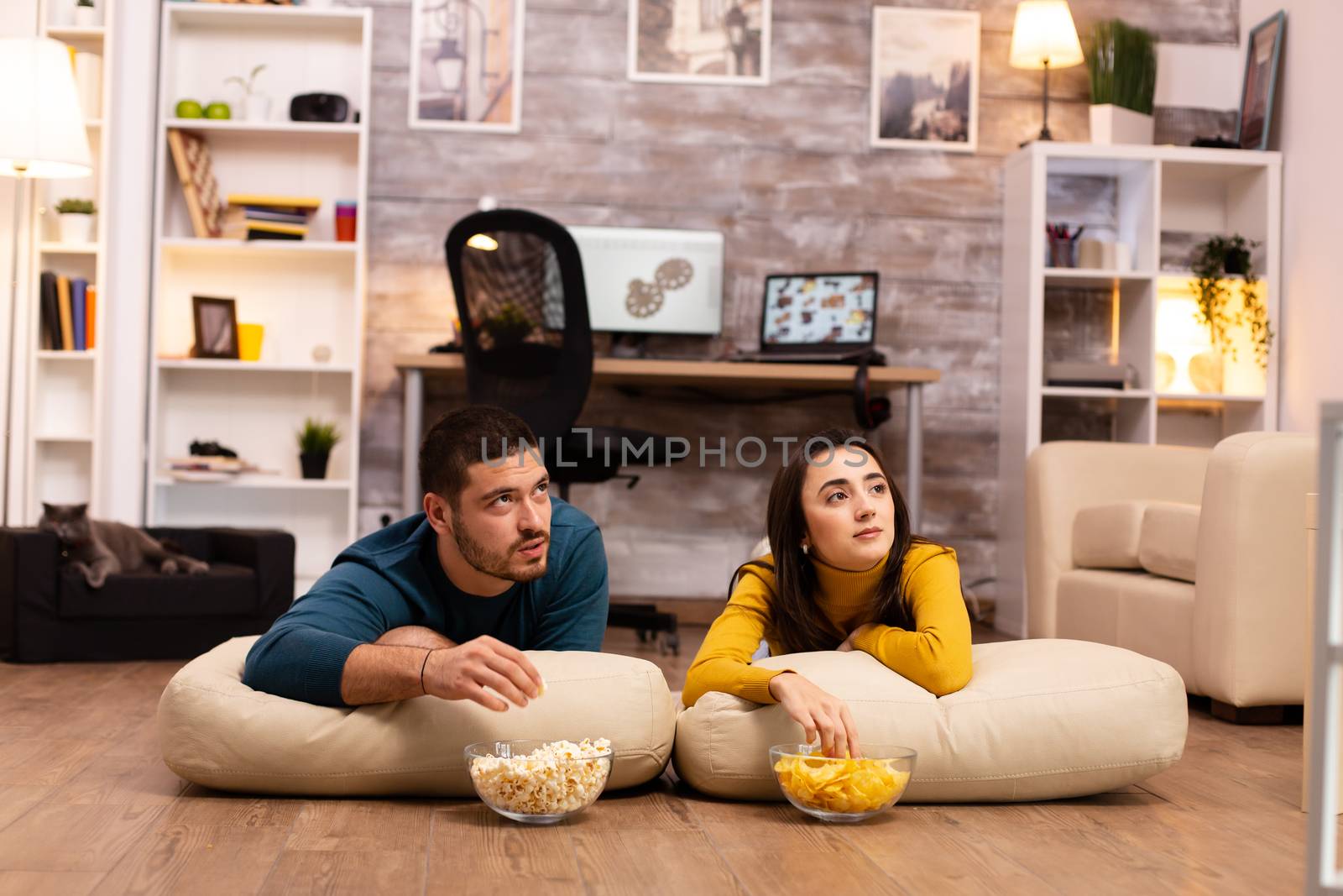 Couple sitting on the floor and watching TV in their living room by DCStudio