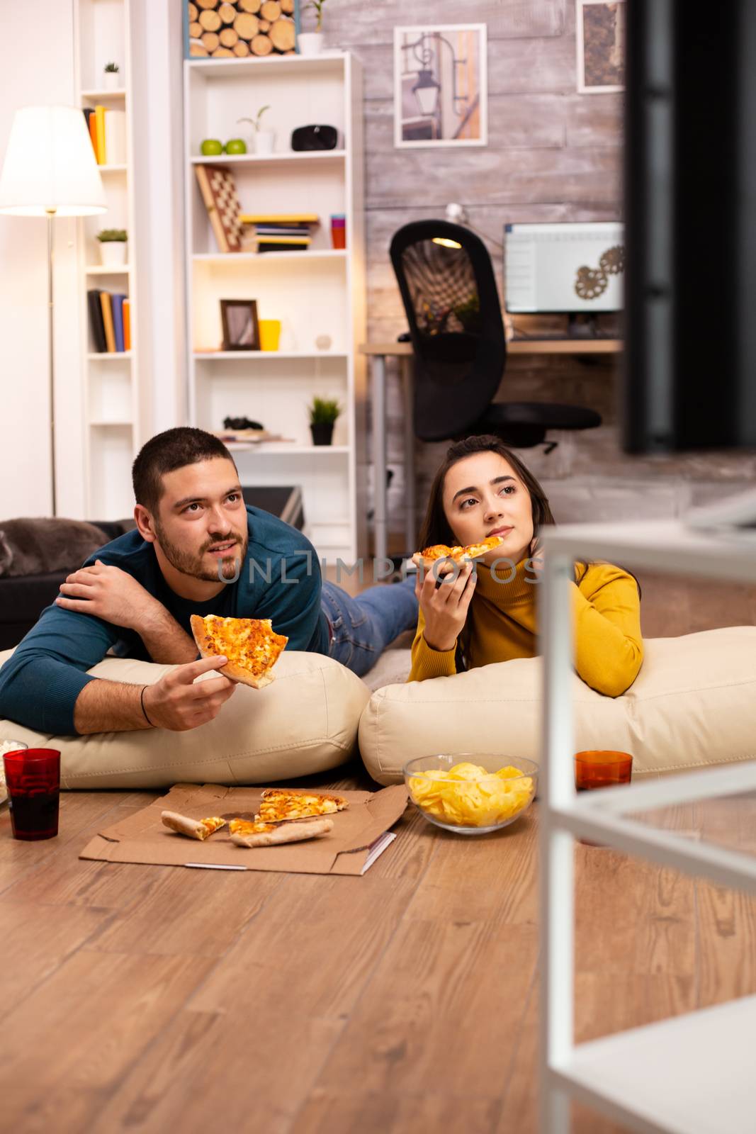 Couple sitting on the floor and watching TV in their living room by DCStudio