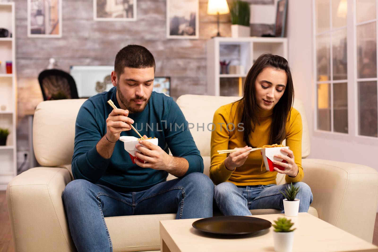 In modern cozy living room couple is enjoying takeaway noodles while watching TV comfortably on the sofa