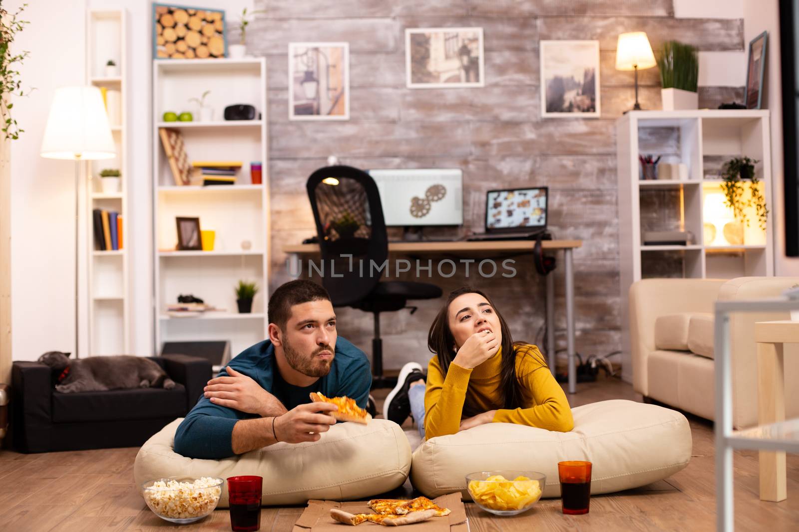 Couple sitting on the floor and watching TV in their living room by DCStudio