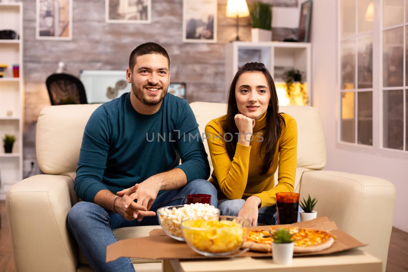 Beautiful young couple watching TV and eating fast food takeaway in the living room