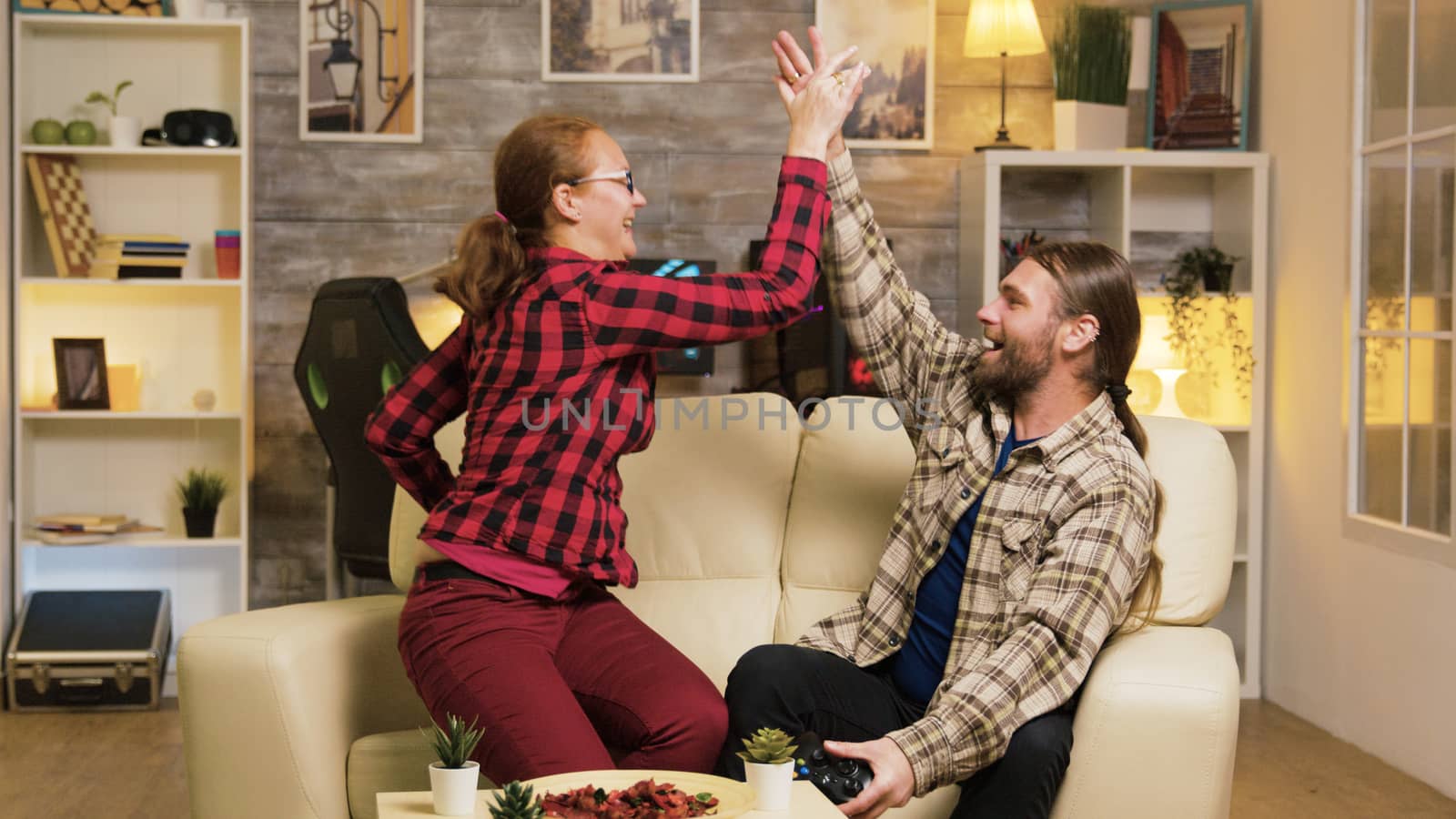 Couple celebrating victory while playing video games using wireless controllers and giving high five.