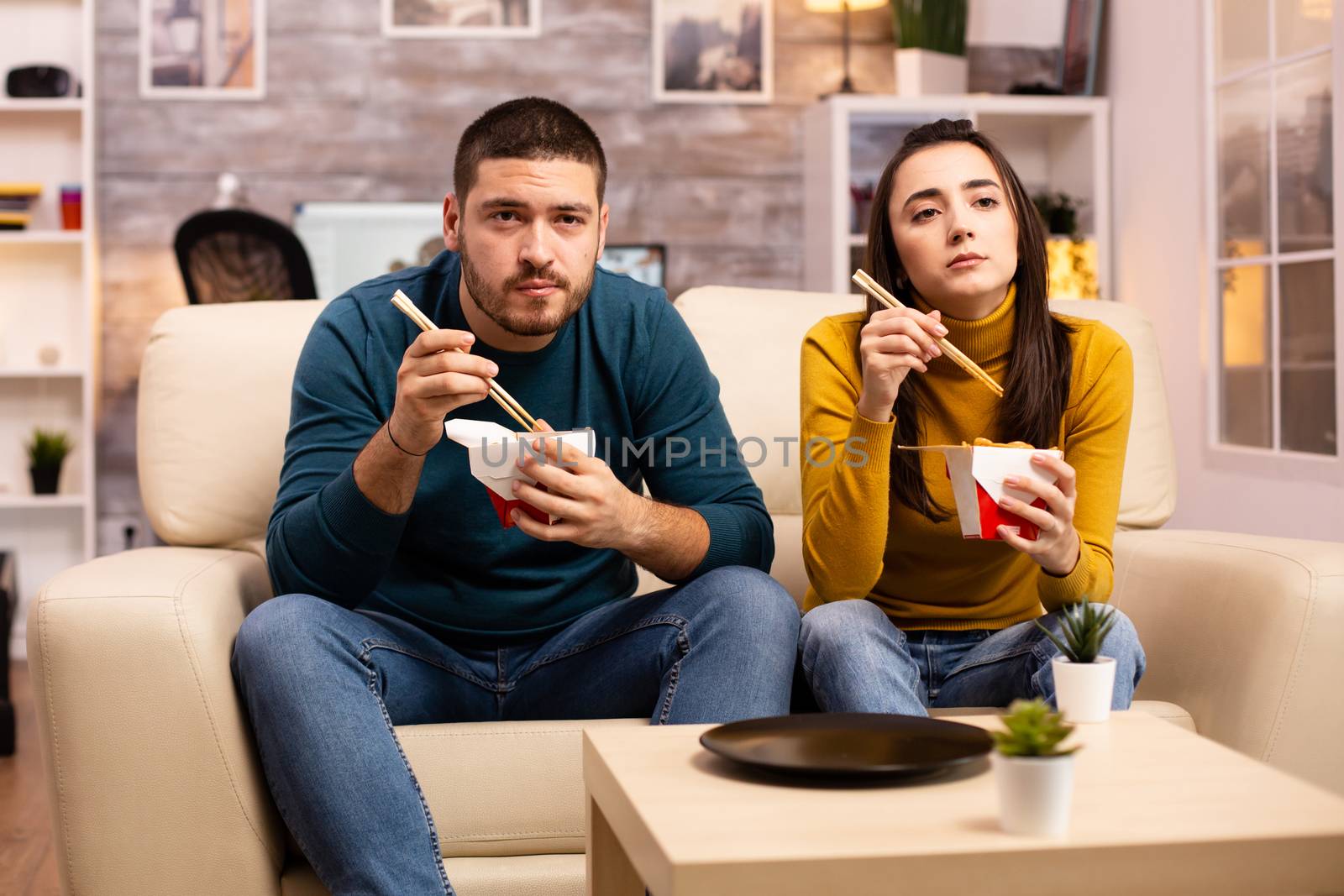 In modern cozy living room couple is enjoying takeaway noodles while watching TV comfortably on the sofa