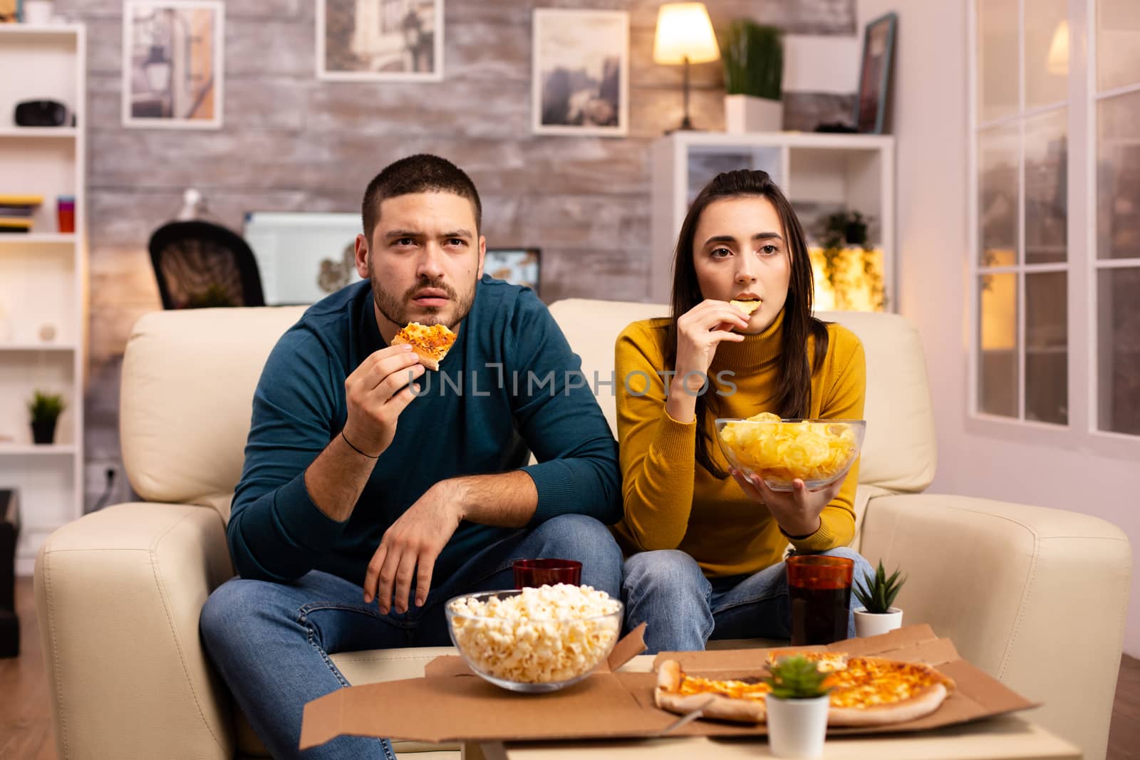 Beautiful young couple watching TV and eating fast food takeaway in the living room