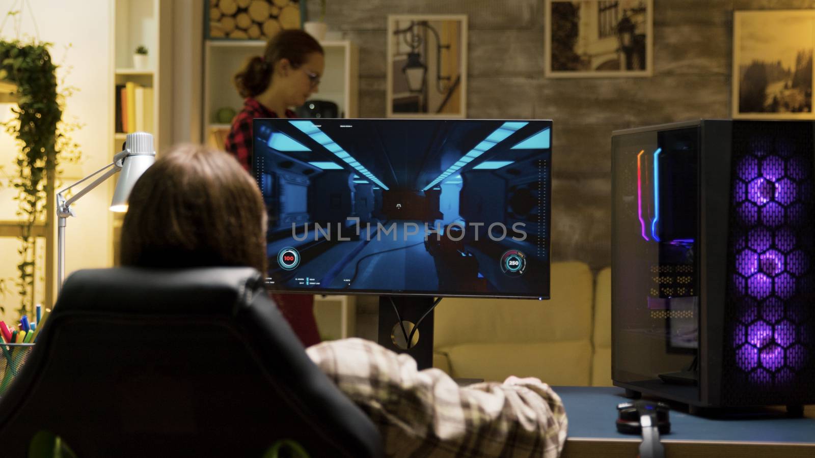 Back view of man sitting on gaming chair and playing games on computer. Girlfriend reading a book in the background.