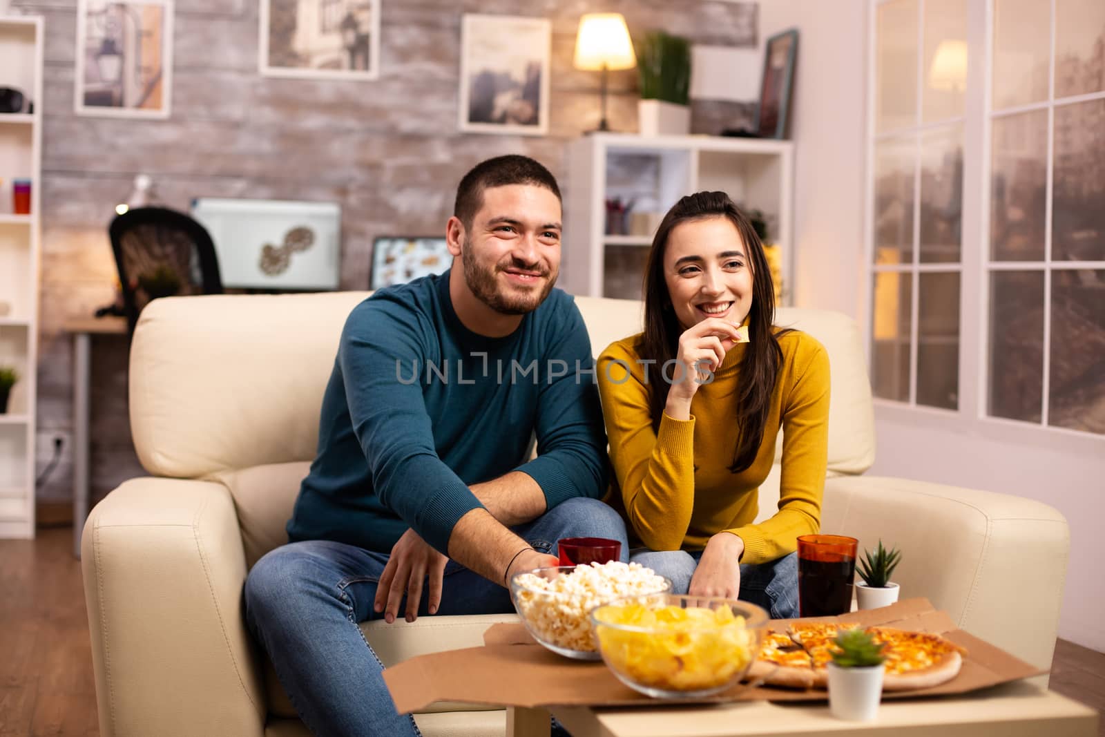 Beautiful young couple watching TV and eating fast food takeaway in the living room