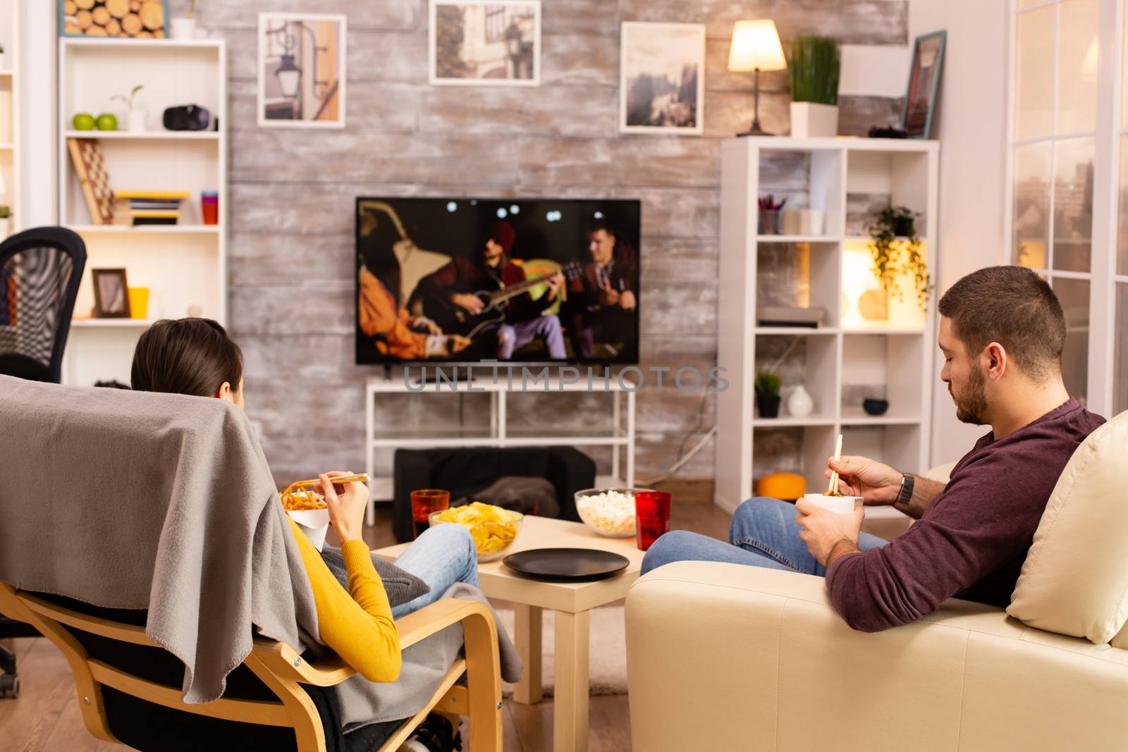 Back view of couple in living room watching a movie on the TV while eating takeaway food