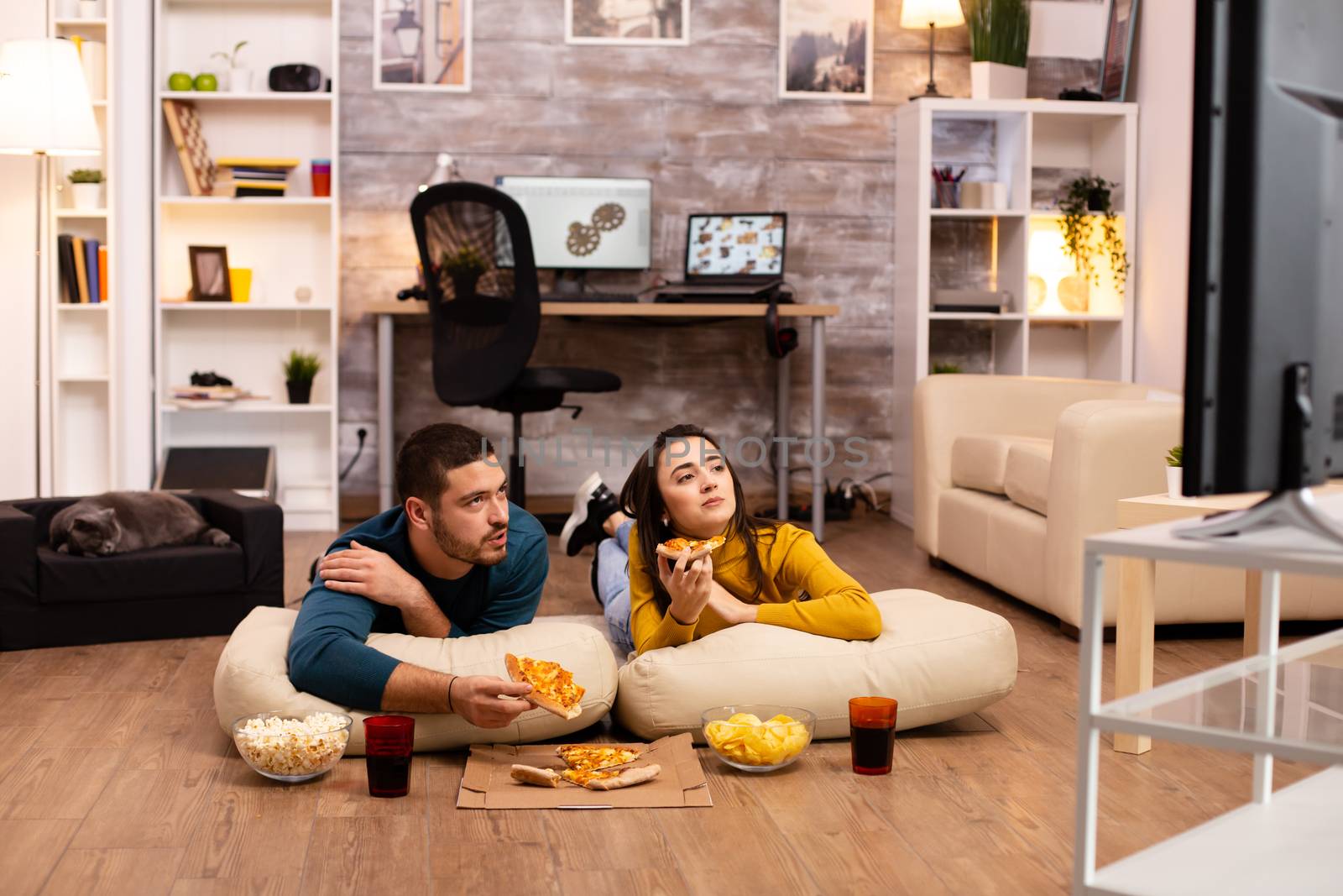 Couple sitting on the floor and watching TV in their living room by DCStudio