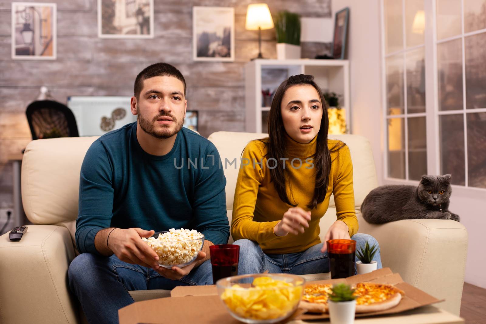 Beautiful young couple watching TV and eating fast food takeaway in the living room