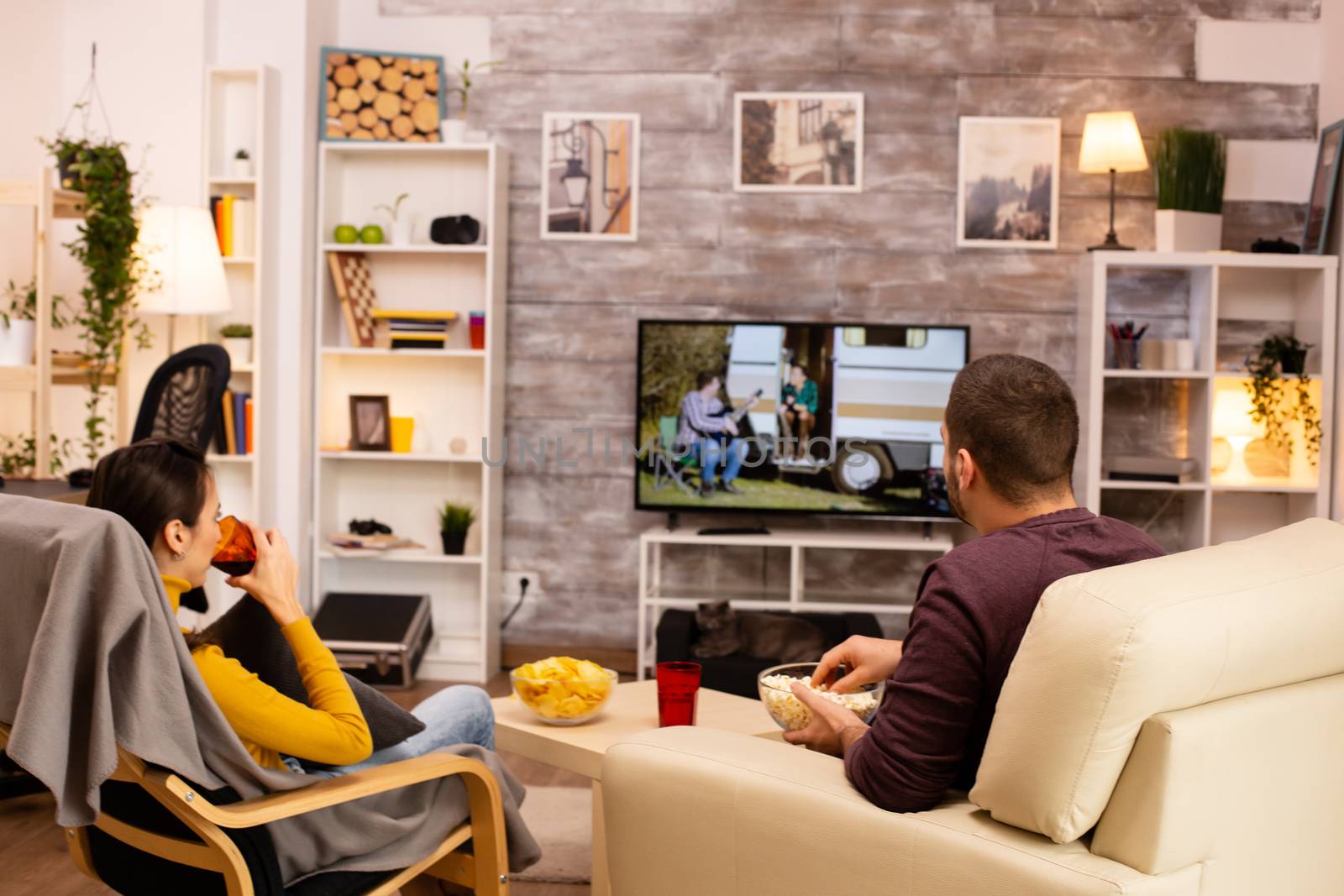 Back view of couple in living room watching a movie on the TV while eating takeaway food