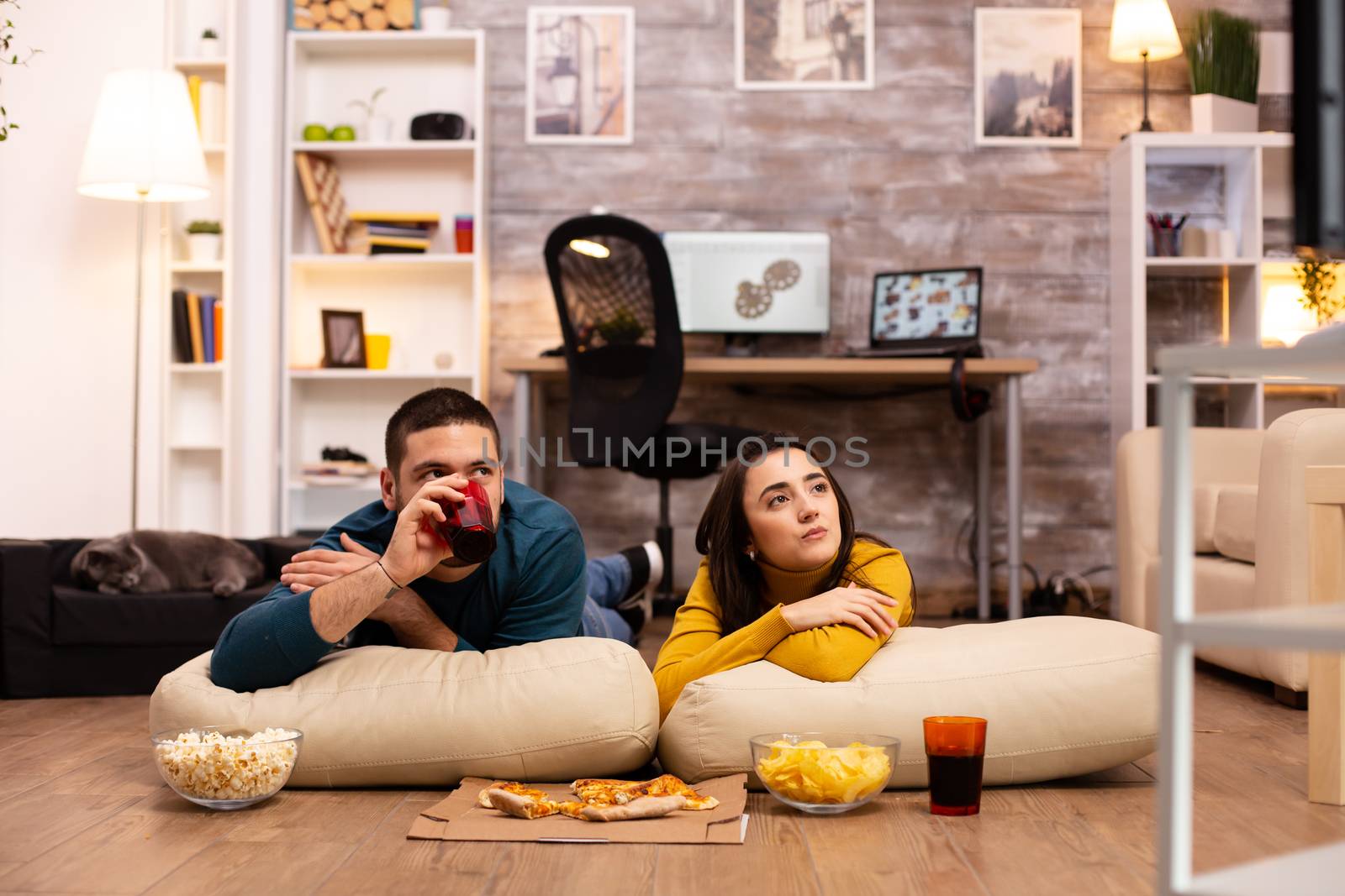 Couple sitting on the floor and watching TV in their living room by DCStudio