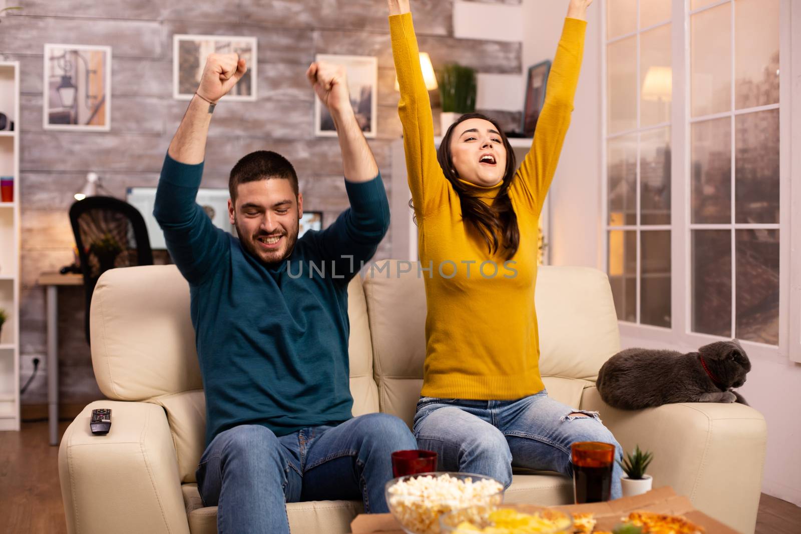 Happy couple cheering for their favourite team while watching TV in he living room by DCStudio