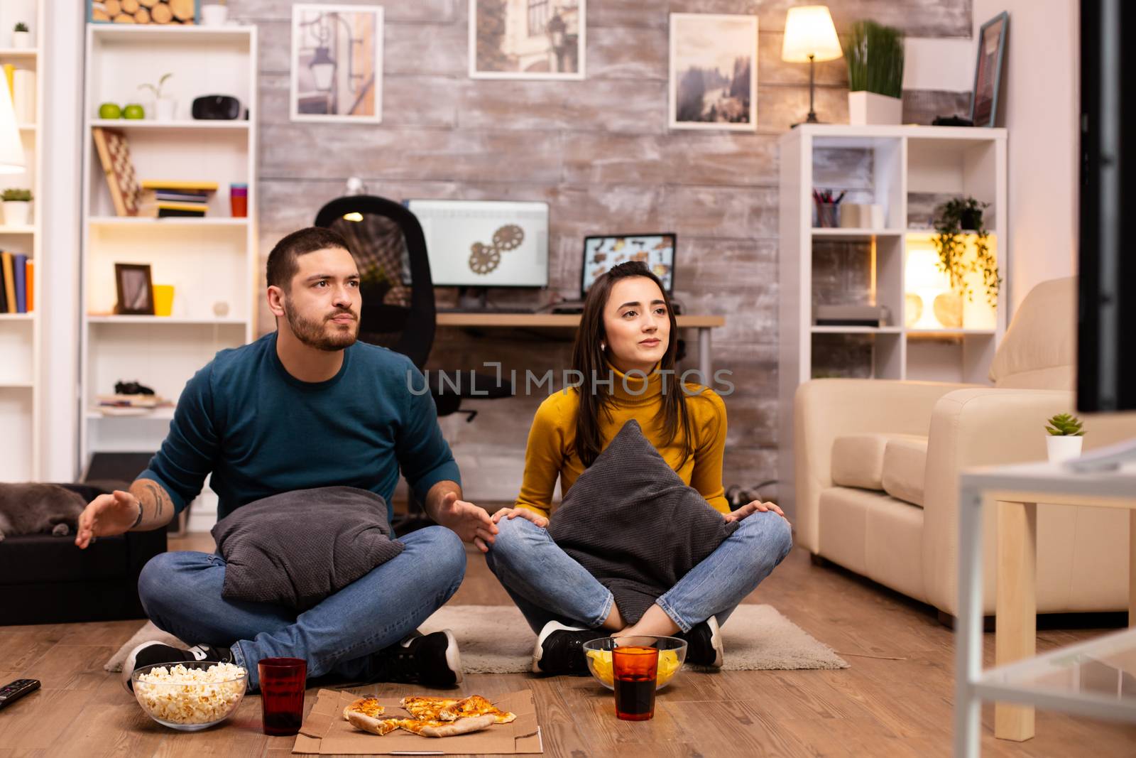 Couple sitting on the floor and watching TV in their living room by DCStudio