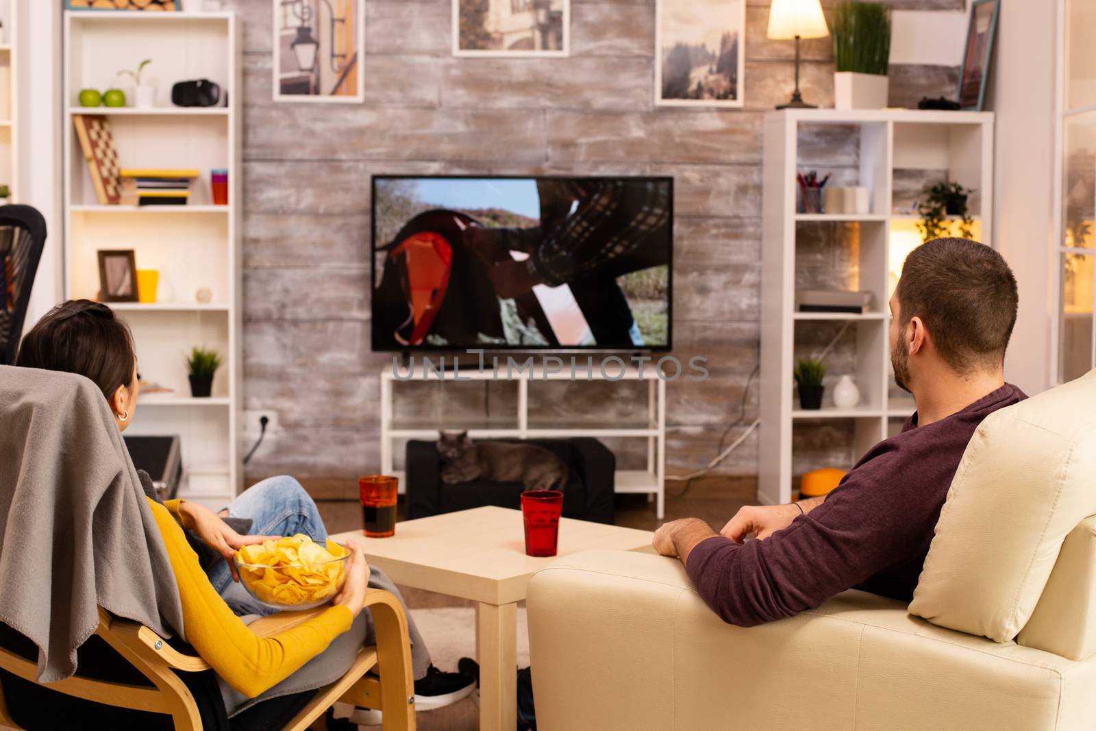 Back view of couple in living room watching a movie on the TV while eating takeaway food