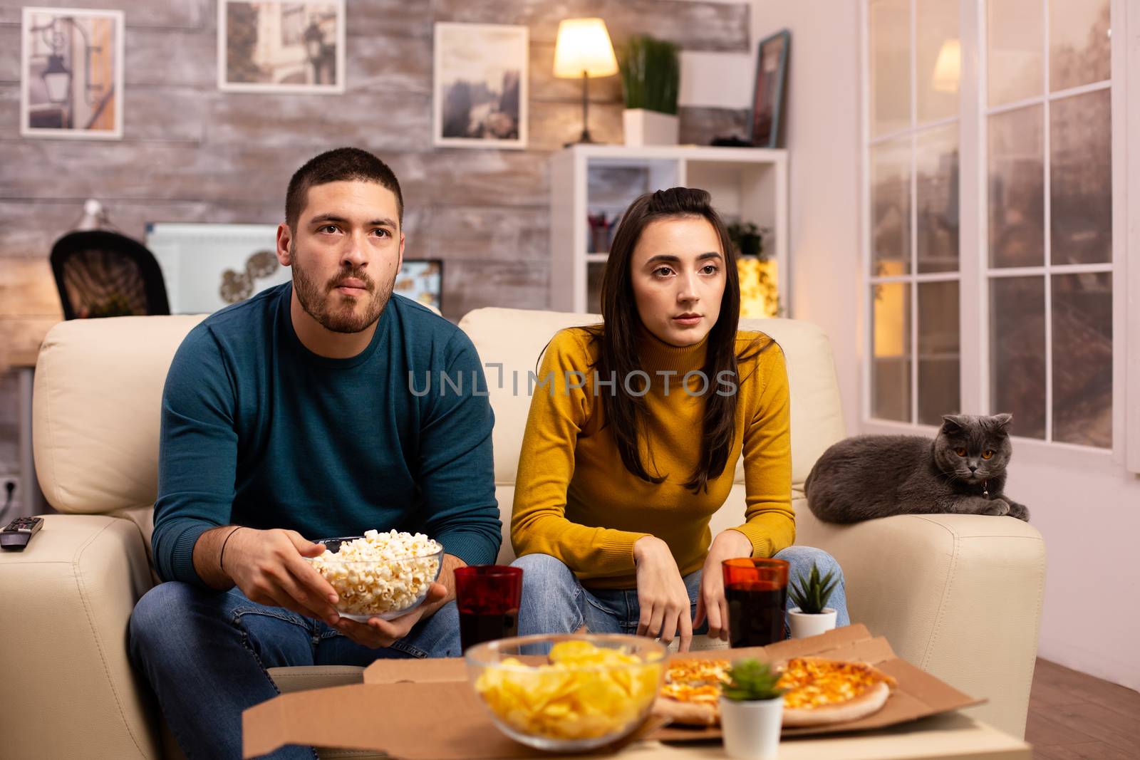 Beautiful young couple watching TV and eating fast food takeaway in the living room