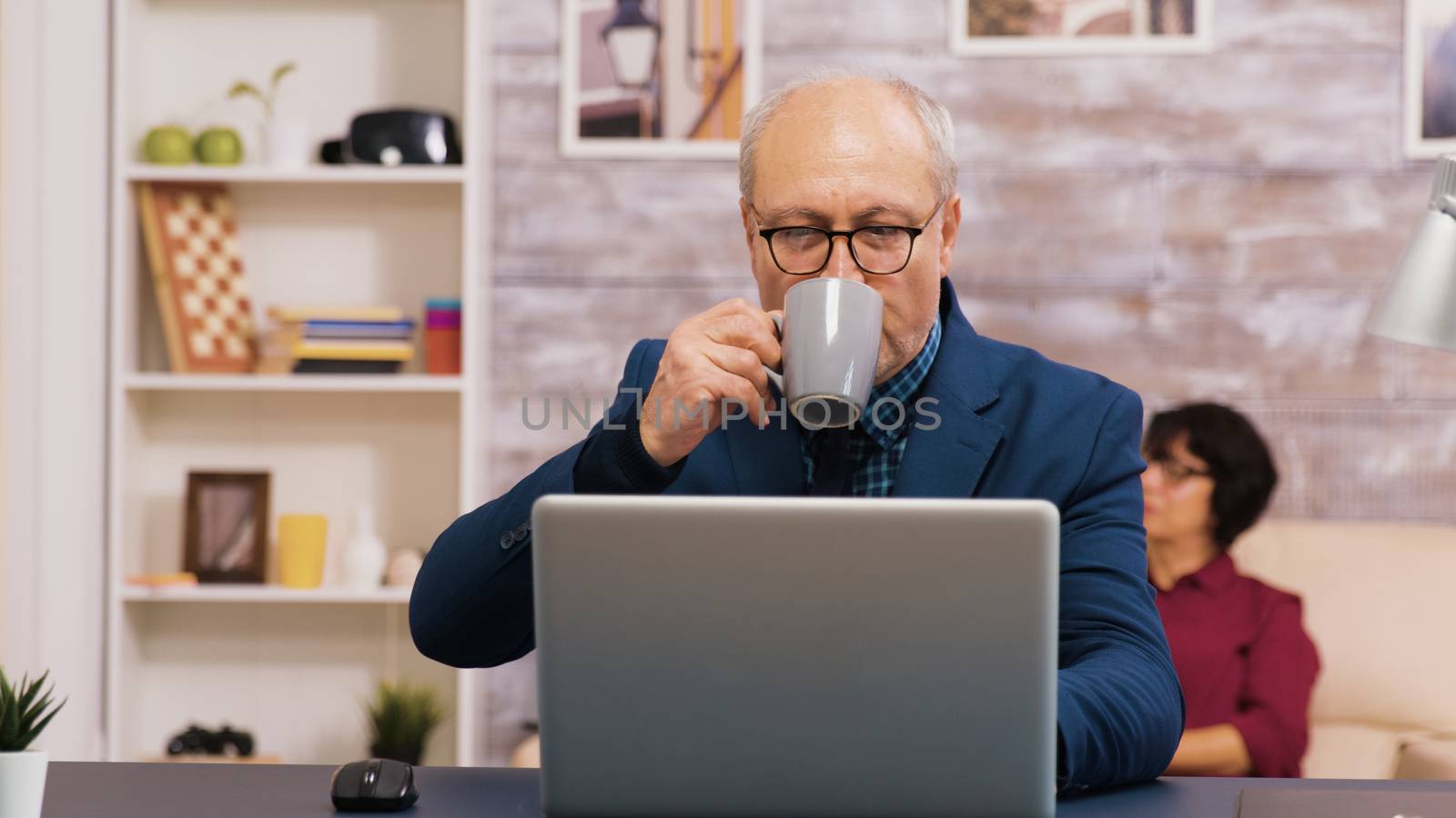 Old man enjoying a cup of coffee while working on laptop in living room. Wife watching tv in the backgroun sitting on the couch.