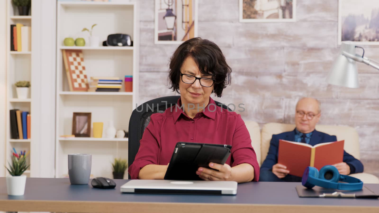 Elderly woman with glasses using tablet in living room. Elederly man sitting on sofa and reading a book in the background.