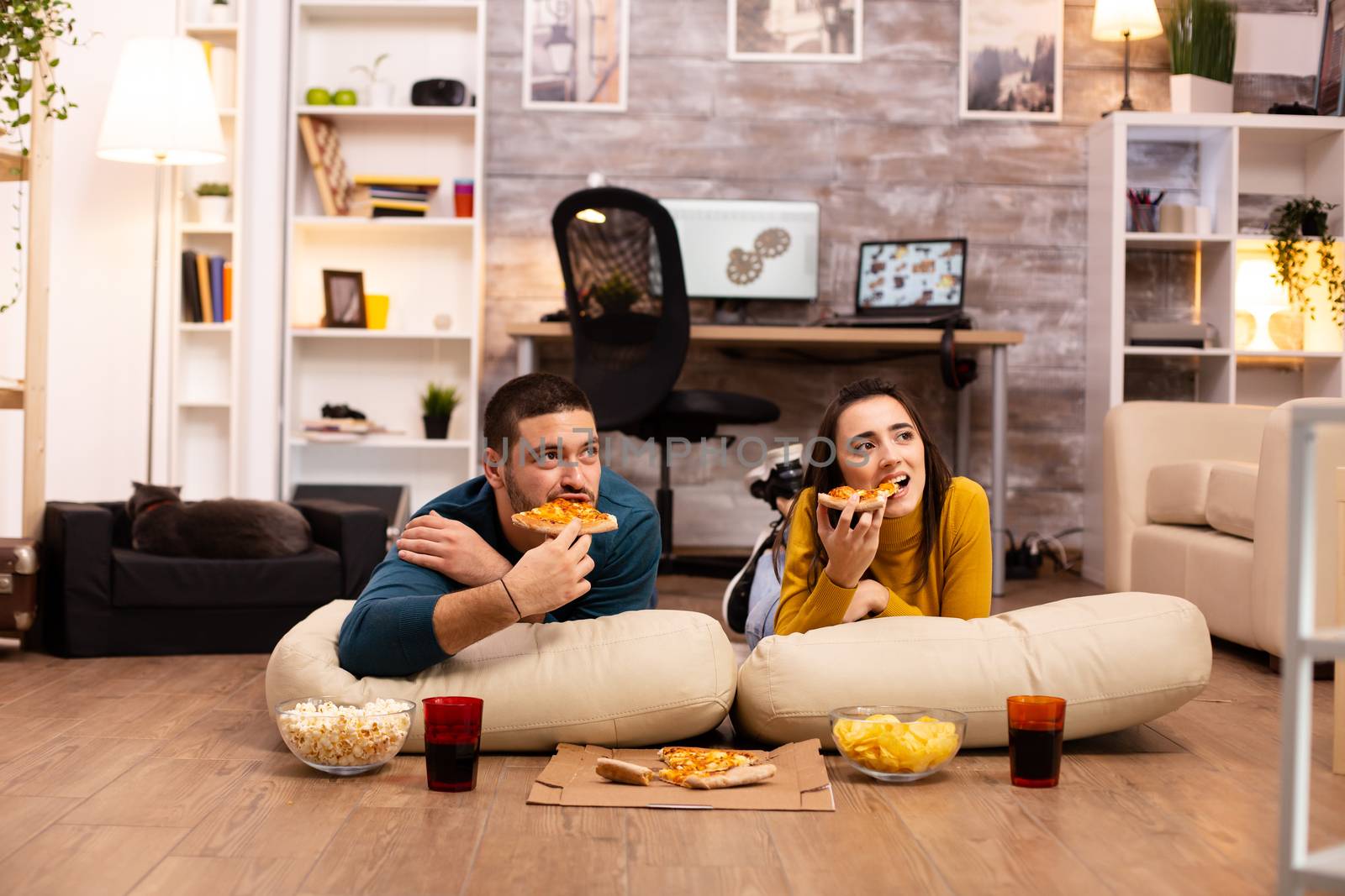 Couple sitting on the floor and watching TV in their living room by DCStudio