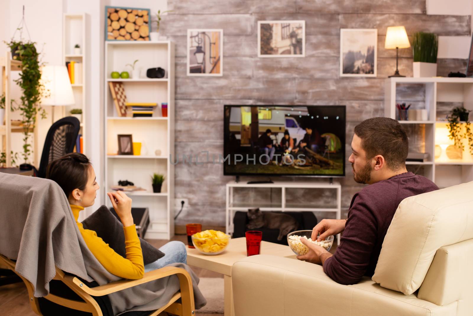 Back view of couple in living room watching a movie on the TV while eating takeaway food