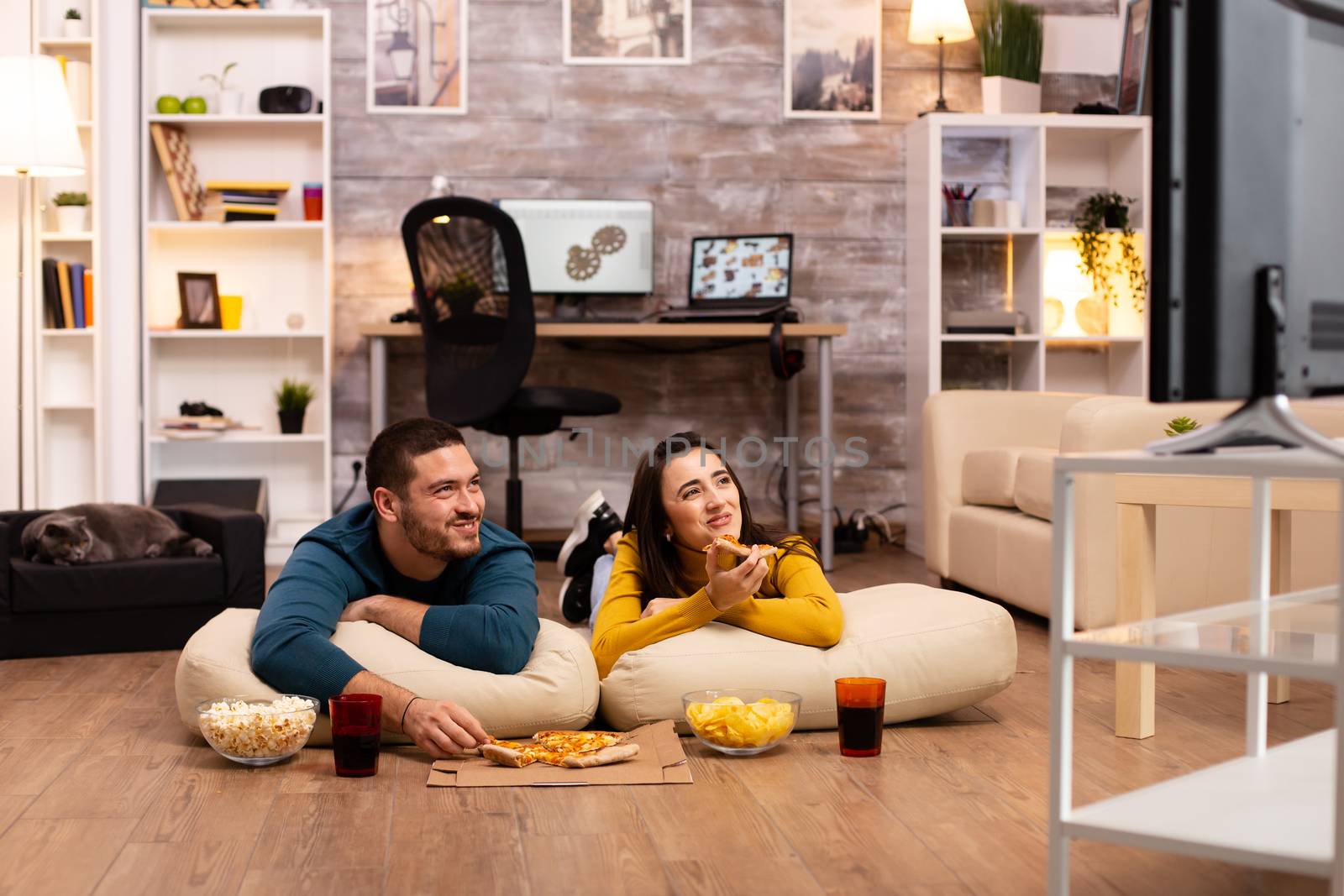 Couple sitting on the floor and watching TV in their living room by DCStudio