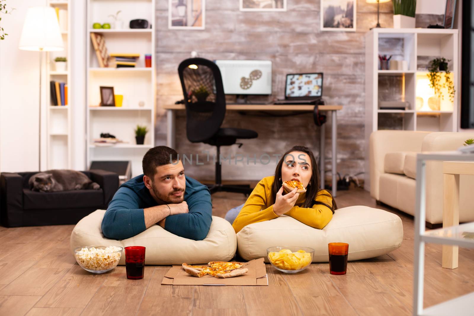 Couple sitting on the floor and watching TV in their living room by DCStudio