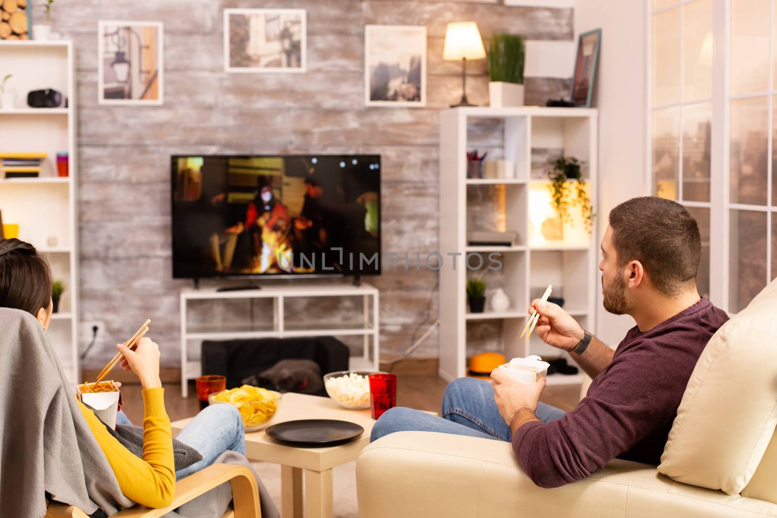 Back view of couple in living room watching a movie on the TV while eating takeaway food