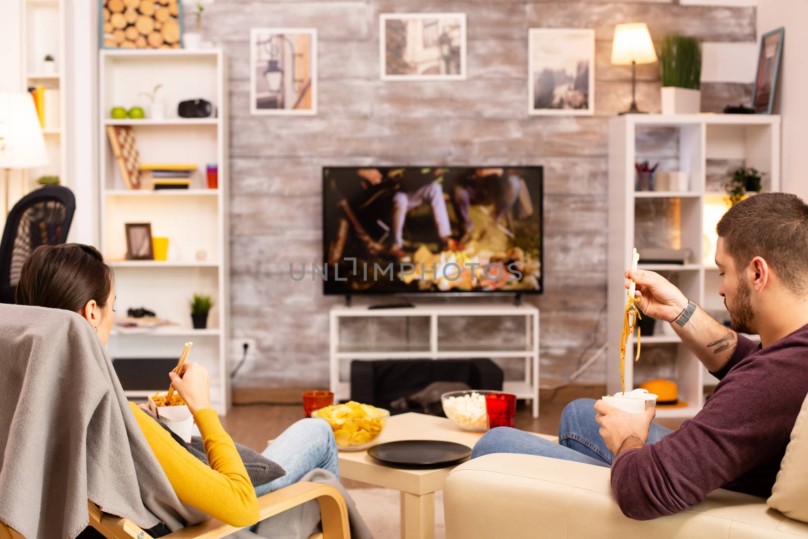 Back view of couple in living room watching a movie on the TV while eating takeaway food