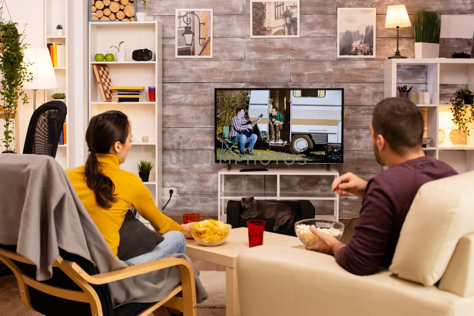 Back view of couple in living room watching a movie on the TV while eating takeaway food