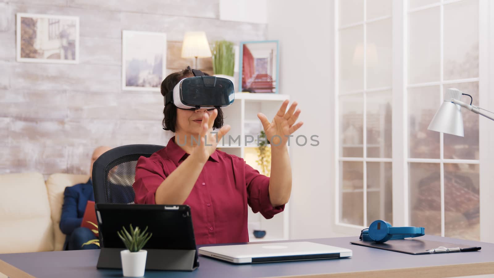 Elderly age woman using virtual reality goggles in living room. Old man sitting on sofa in the background.